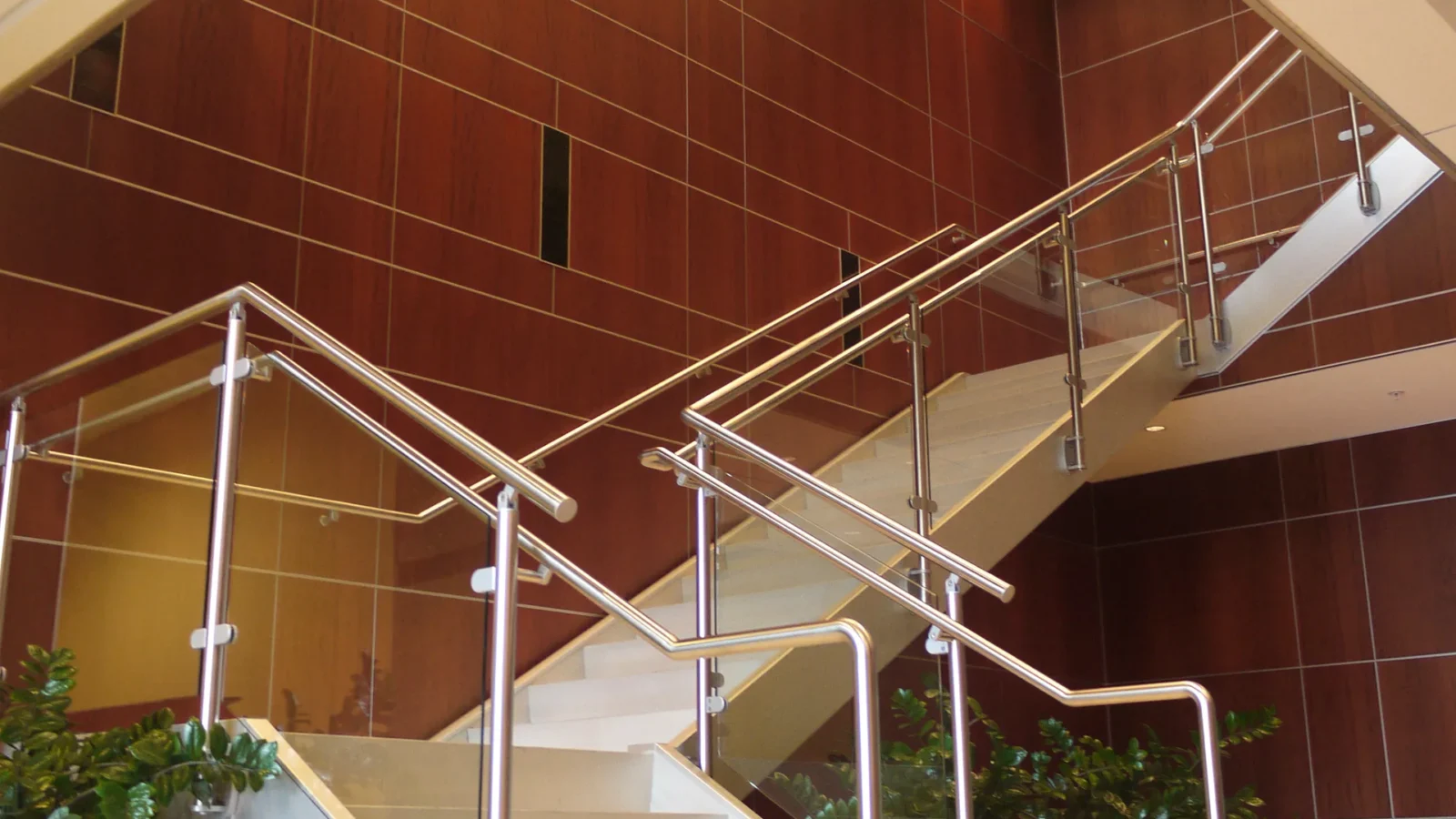 Lobby stair with stainless steel handrails and glass guards rising in front of a tall wall of cherry wood panels.