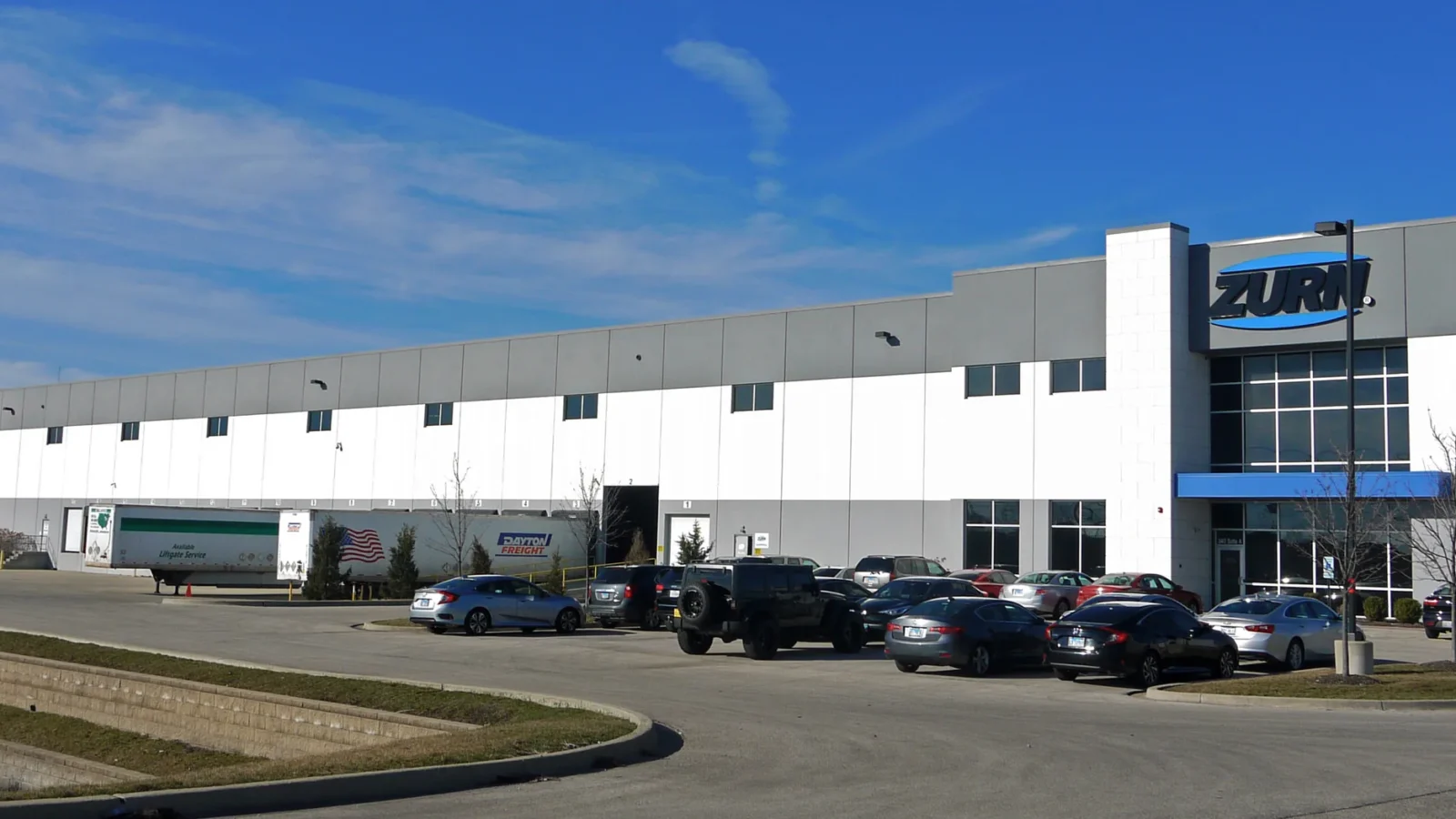 Full length of the multi-tenant warehouse from across the parking lot, showing the Zurn office end and the loading docks beyond.