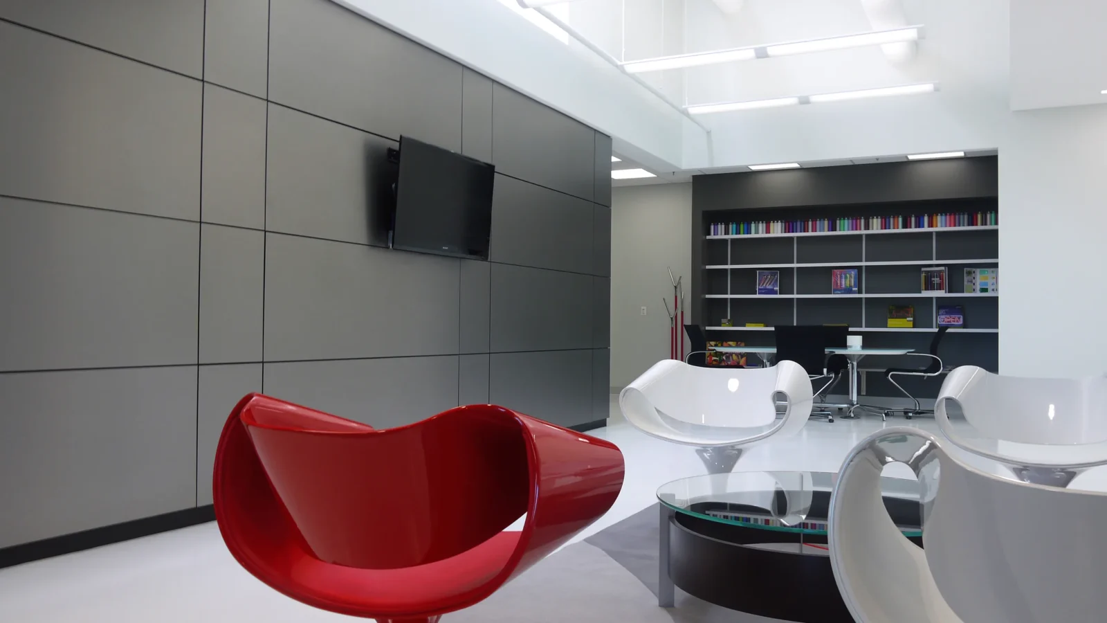 Welcome lounge with a glossy red sculptural chair, white swivel seats around a glass table, grey panelled wall and a color-sample library behind.