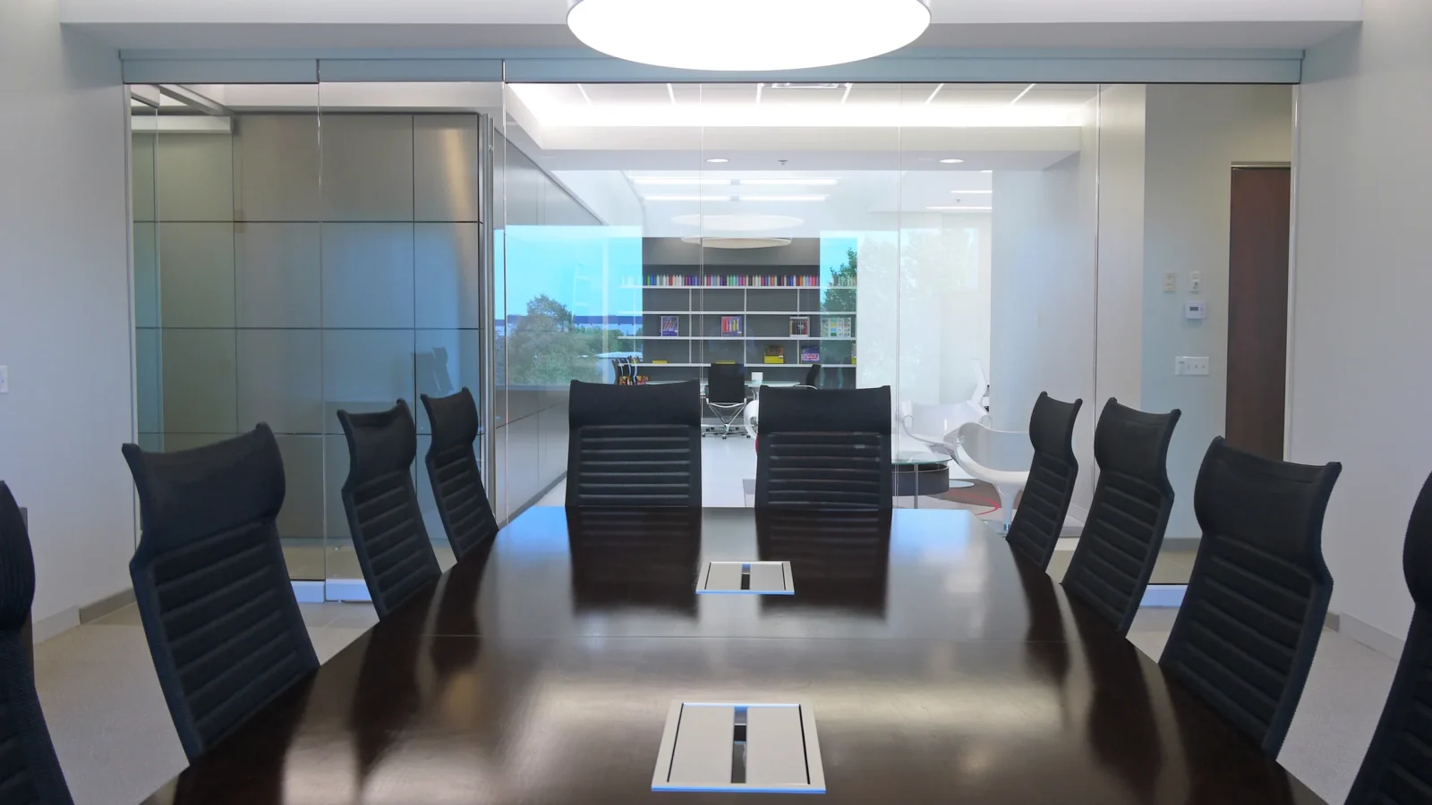 Glass-walled conference room with a dark wood table, black mesh chairs and a round pendant light, looking through to the studio.