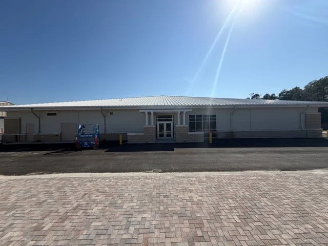 The long, low front elevation of the Eglin AFB test facility: light metal panel walls on a masonry base, a standing-seam roof and a paved plaza before the entry.