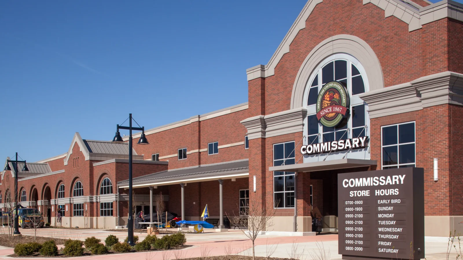The Fort Belvoir Commissary in daylight: a red-brick front with a tall arched entry bay carrying the Commissary medallion, cream trim, and a covered walkway along the storefront.