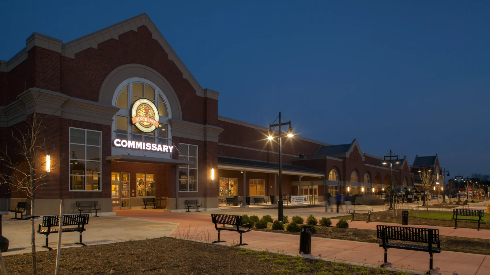 The Fort Belvoir Commissary at dusk, its lit arched entry and Commissary sign glowing against a deep blue sky, with benches and lamp posts along the entry plaza.