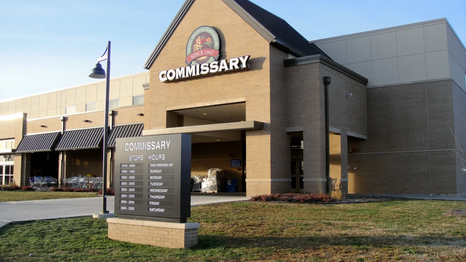 Brick gabled entrance with the Commissary sign and DeCA emblem above a covered entry, with a brick-based store hours monument sign in front.