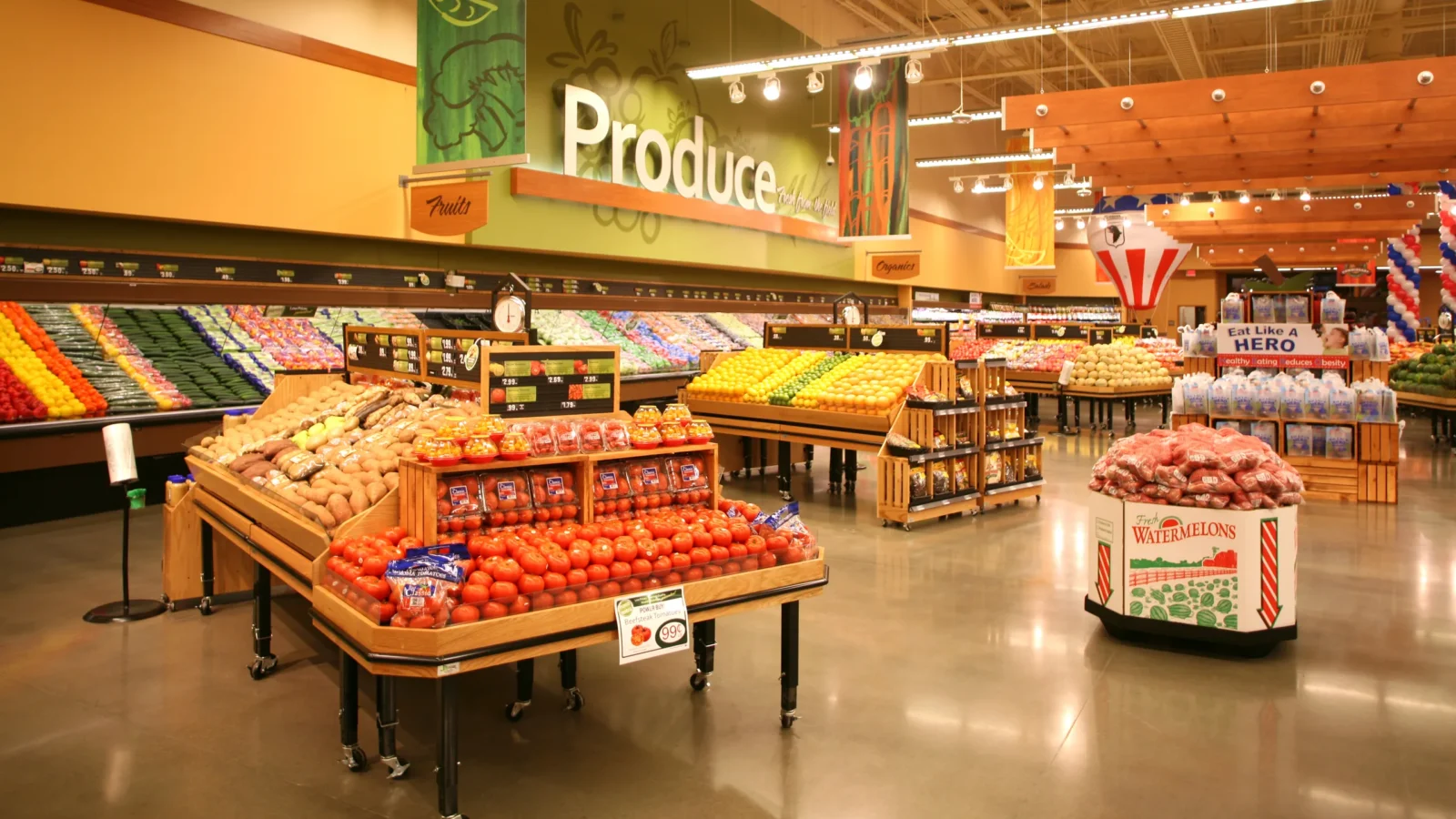 Produce department with green Produce wall graphic, wood display tables of tomatoes and citrus, and warm wood ceiling clouds over polished concrete.