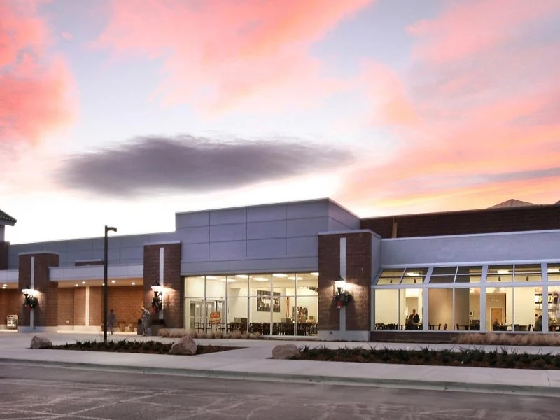 The Fort Carson Exchange at dusk — the full storefront under a pink sunset sky, with the gabled Exchange entry at left and a lit, glazed retail frontage running to the right.