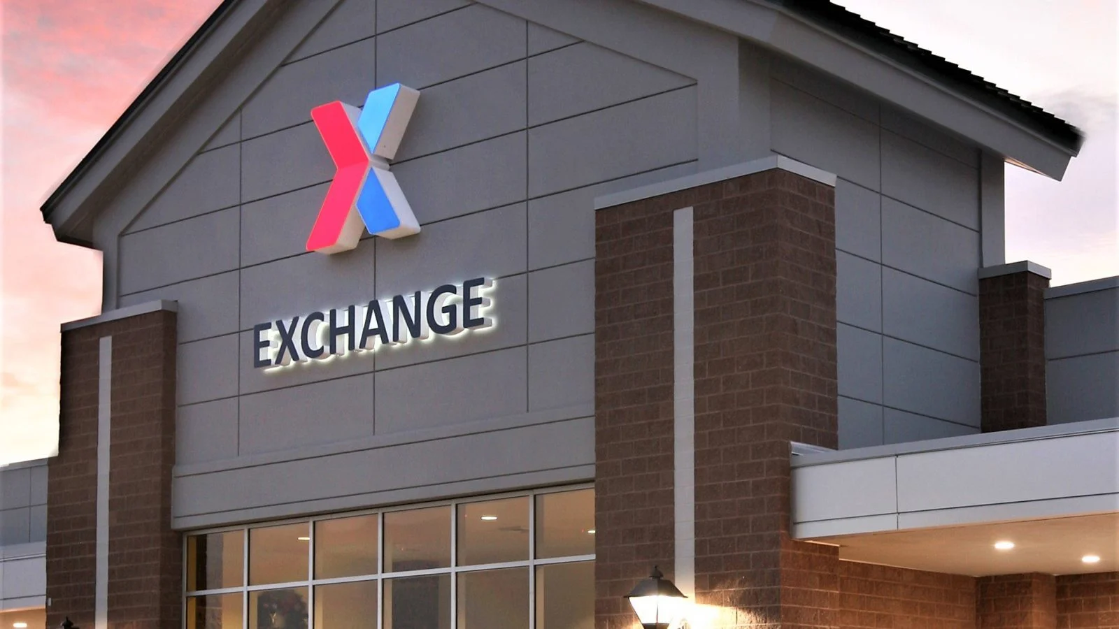 The Fort Carson Exchange entry at dusk — a gabled grey-panelled tower carrying the AAFES X mark and illuminated EXCHANGE lettering above glazed doors, flanked by brick piers.