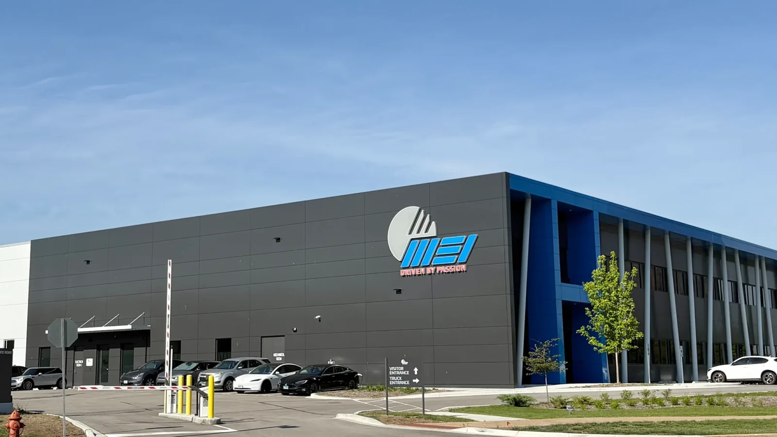Office block and warehouse volume with the illuminated blue MEI logo, flagpoles, monument sign and visitor and truck entrance drive.