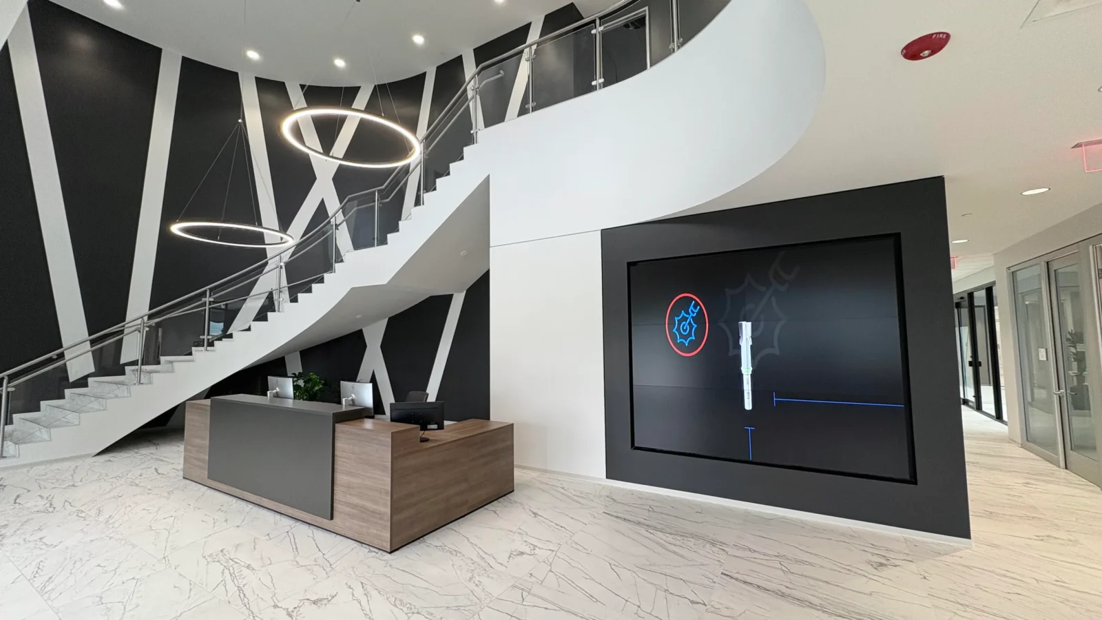 Lobby with a sweeping white stair, ring pendant lights, black and white graphic wall, wood reception desk and marble-look floor.