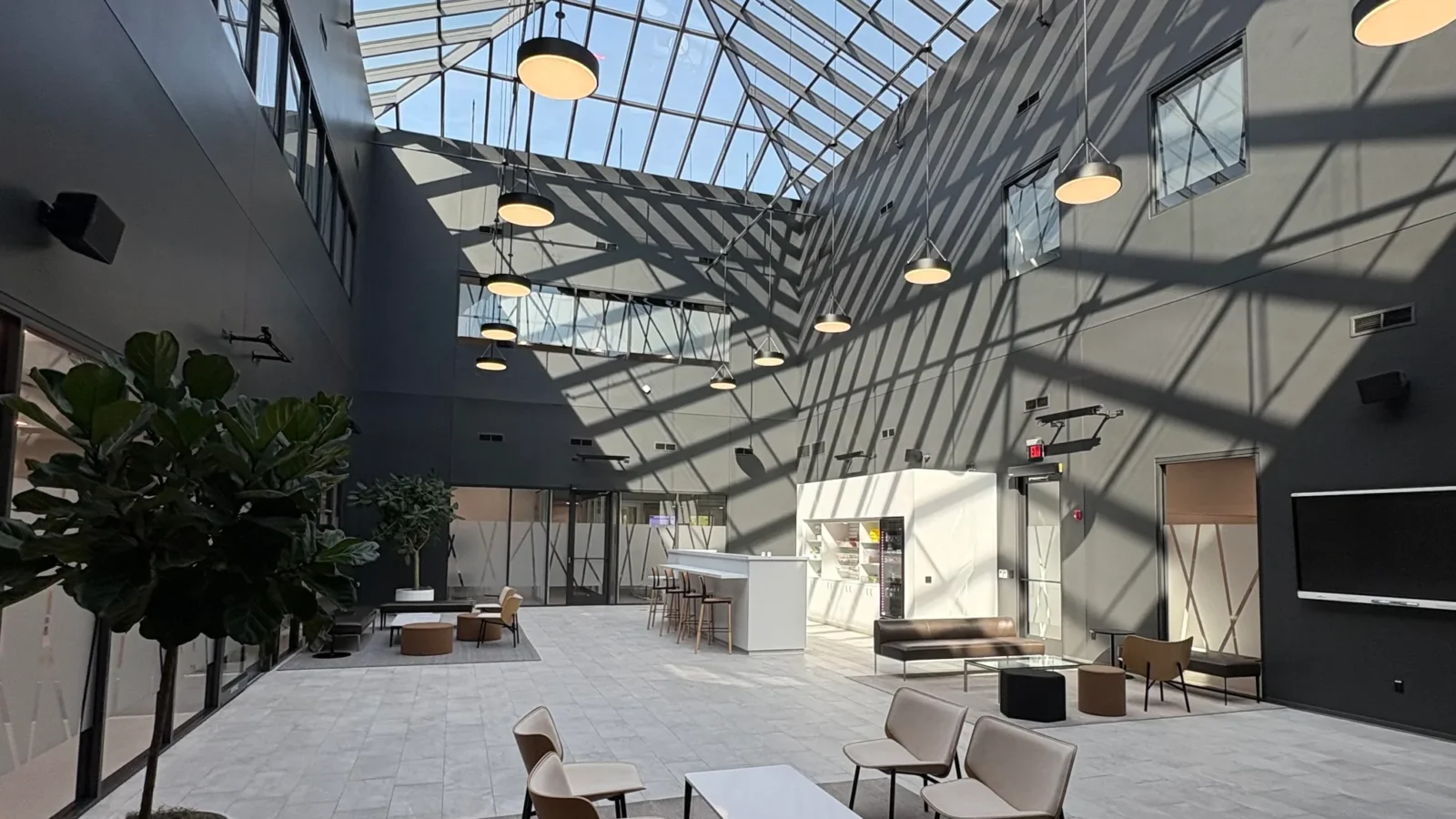 Two-story atrium under a pitched glass skylight, with lounge seating, planters and a coffee bar casting sharp shadow patterns.