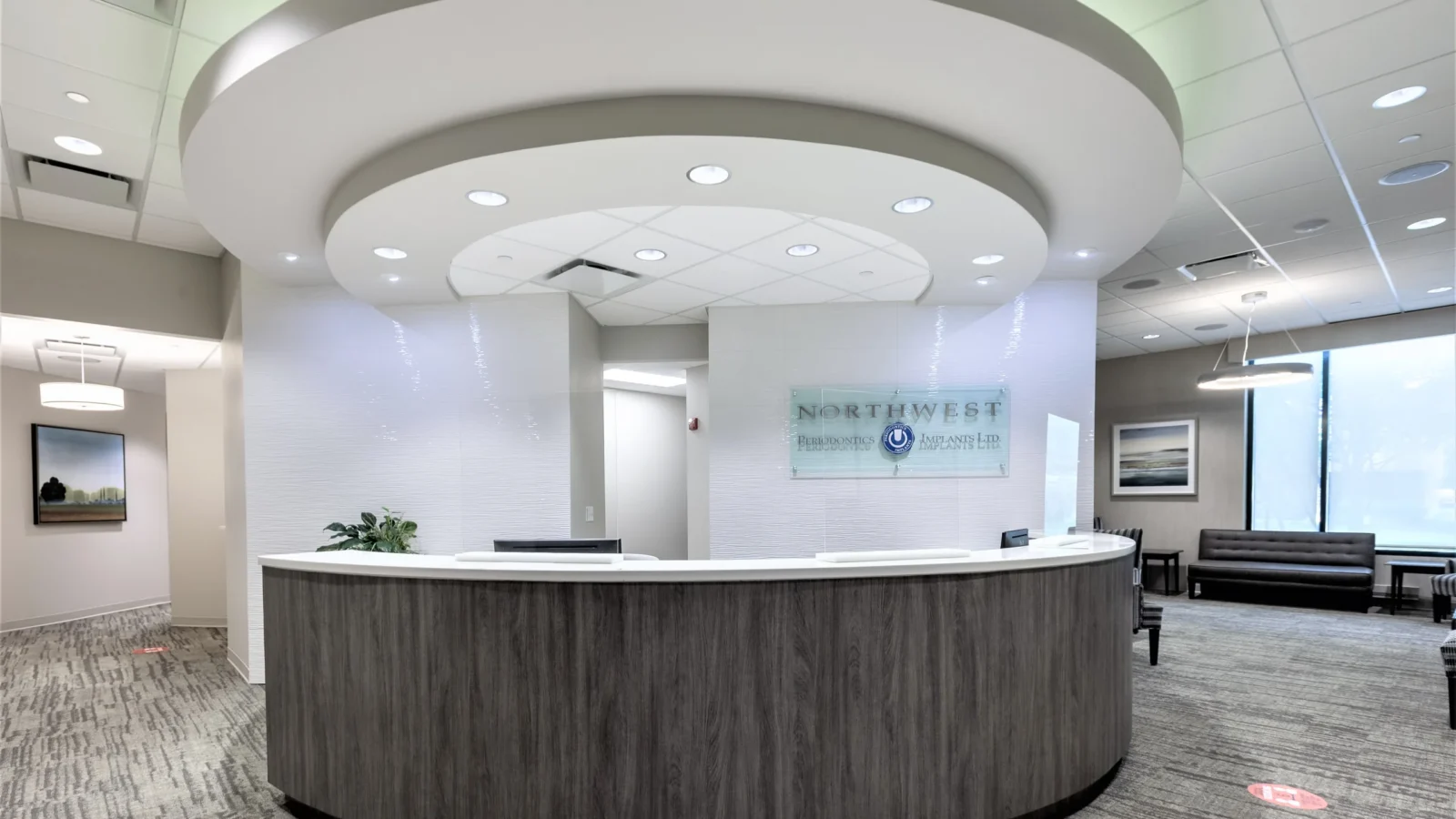 Northwest Periodontics Office reception, a curved wood-grain desk under a tiered circular ceiling with glass logo signage and waiting area beyond.