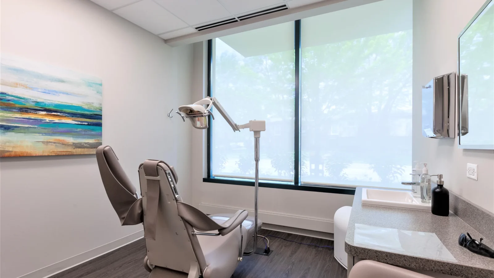 Daylit treatment room with a dental chair, solid-surface counter and sink, wood-look plank floor and a coastal painting on the wall.