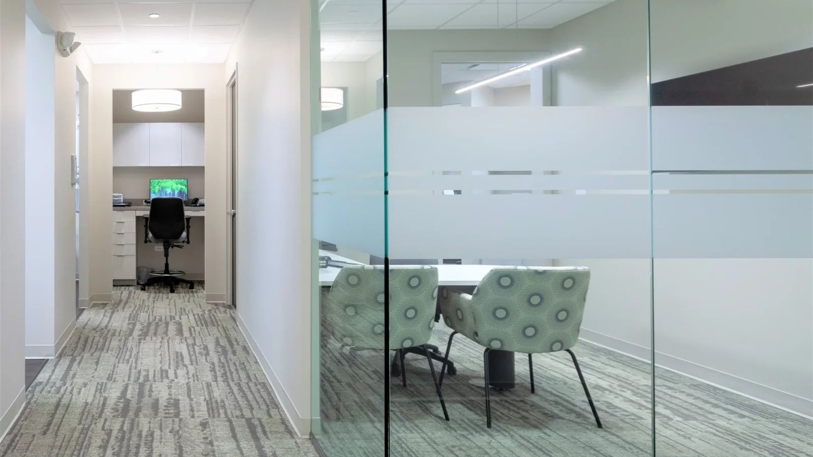 Clinical corridor alongside a glass-walled consultation room with frosted banding, patterned chairs and textured carpet.