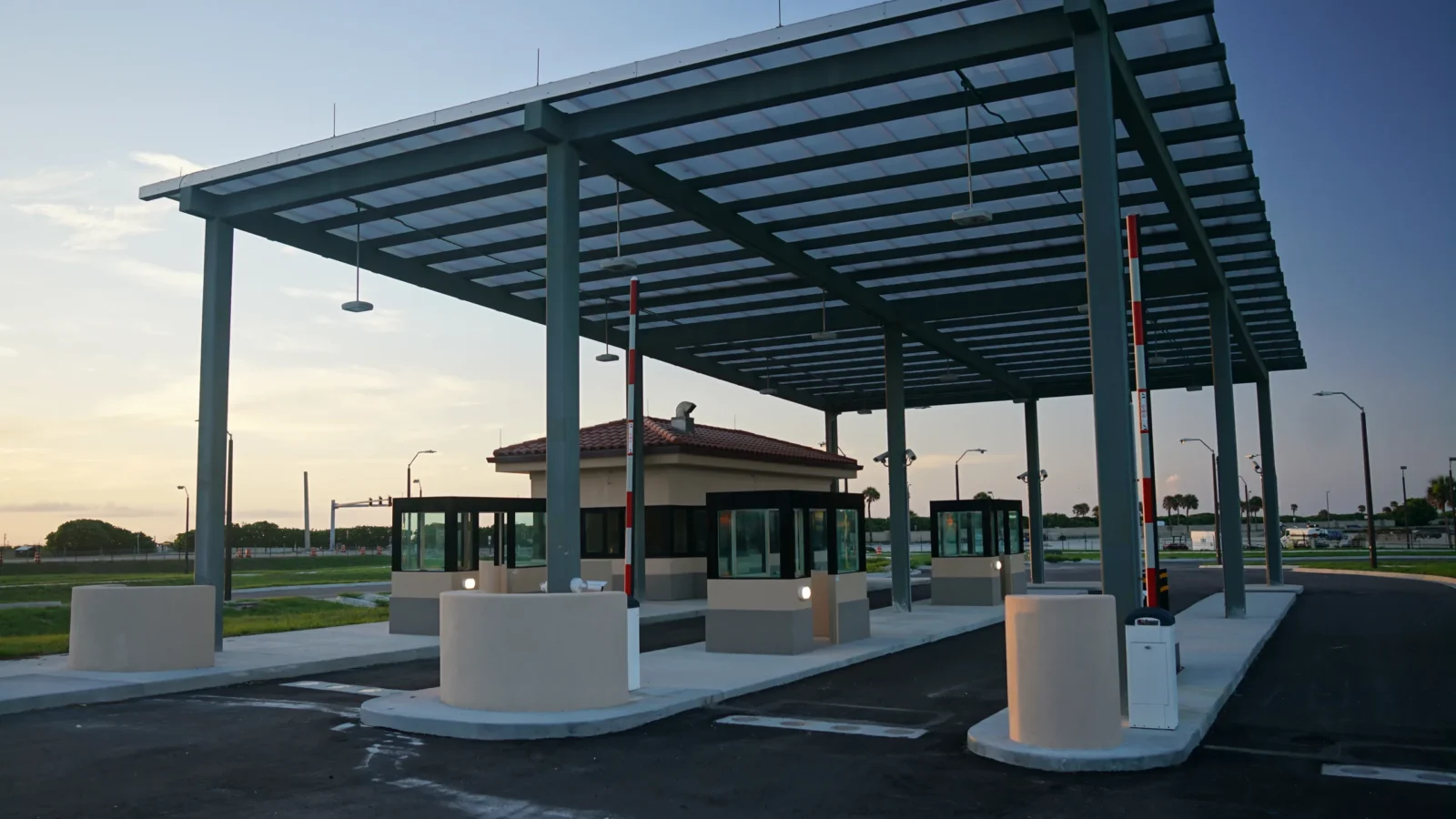 Patrick SFB Gate at dusk: steel inspection canopy over guard booths and stucco barrier islands, with the tile-roofed gatehouse behind.