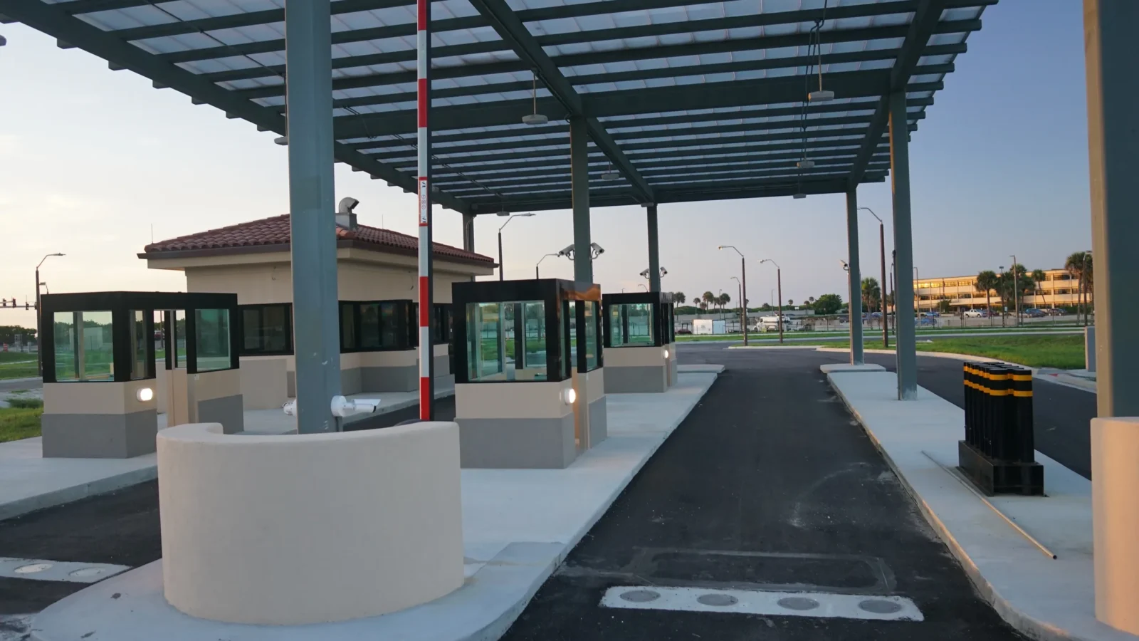 Under the inspection canopy at dusk: curved stucco barrier islands and glass-walled guard booths line the lanes, with a red-and-white barrier arm and the tile-roofed gatehouse beyond.