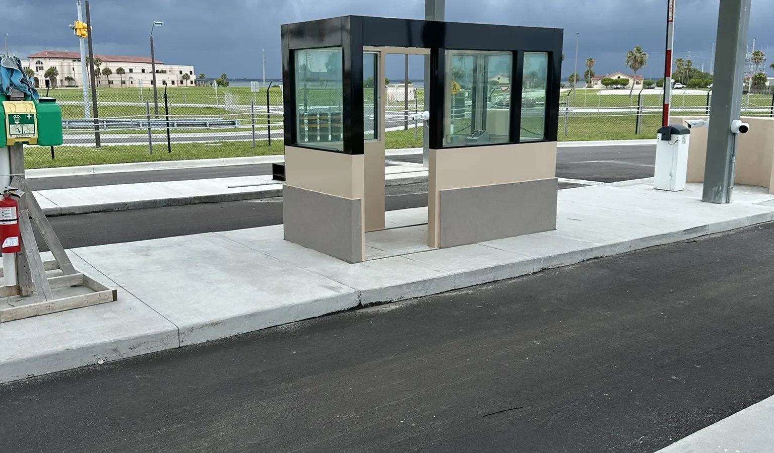 A glass-walled guard booth on its island beneath the steel inspection canopy, with a lane barrier arm beside it under a stormy sky.