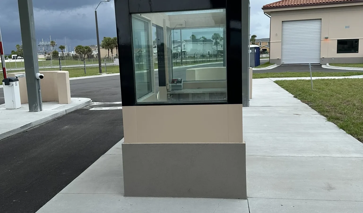 A guard booth seen along the island walkway: dark-framed glazing over a two-tone stucco base, with the gatehouse beyond.