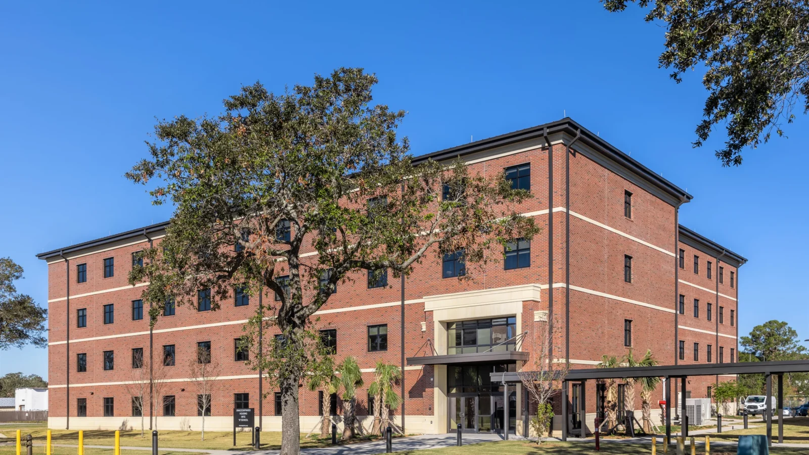 Residence Hall P723 at Corry Station, a four-story red brick residence hall with cast-stone bands and a covered walkway, behind a mature live oak.