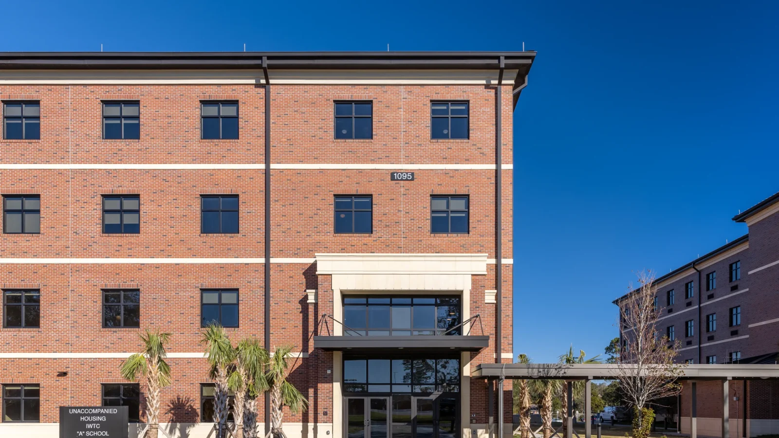 Main entry of Building 1095 with a two-story glazed entrance, cast-stone surround, steel canopy, palms and the Unaccompanied Housing sign.