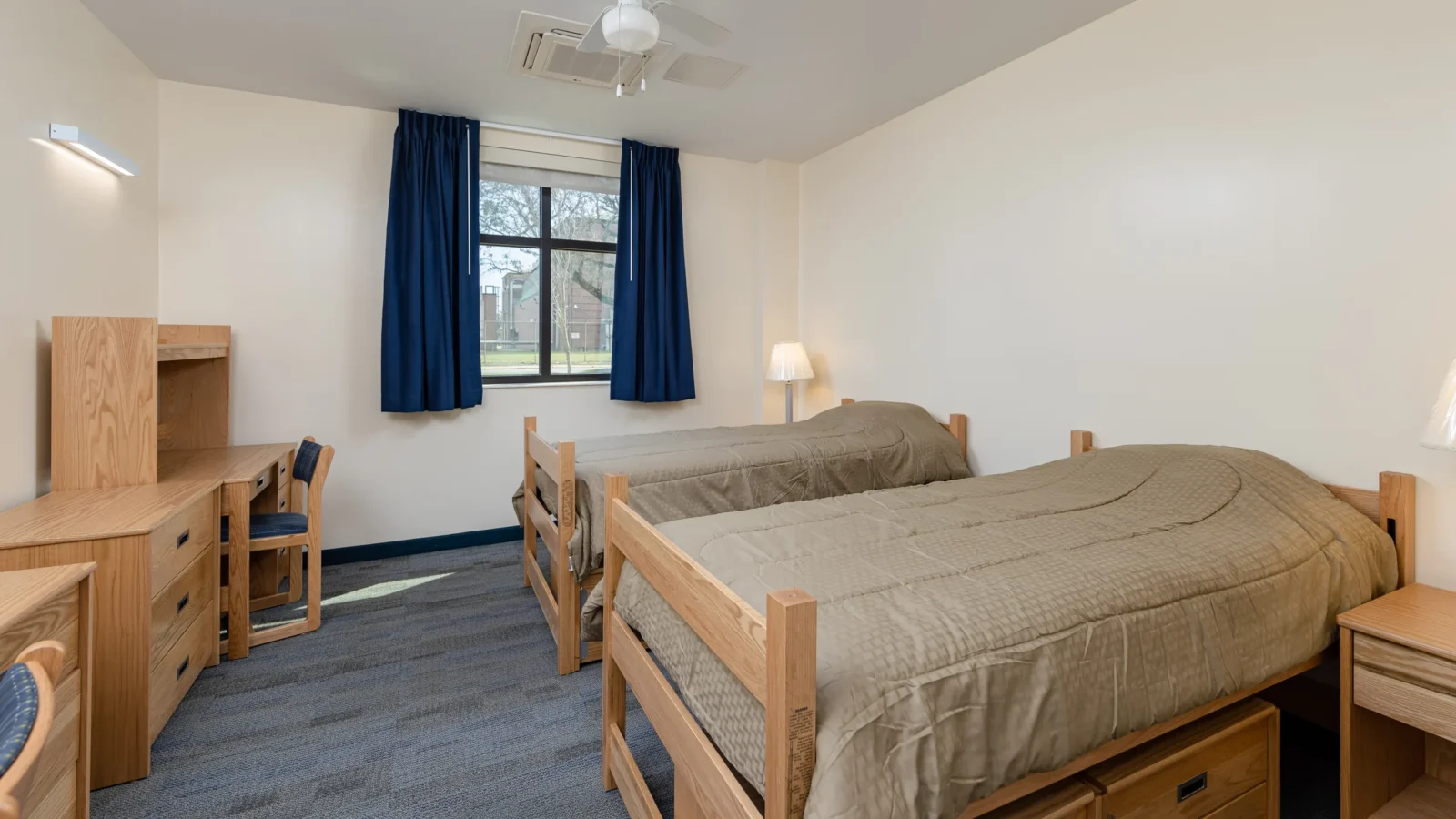 Two-person living quarters with oak beds, desks and dressers, blue carpet tile and a daylit window framed by navy curtains.