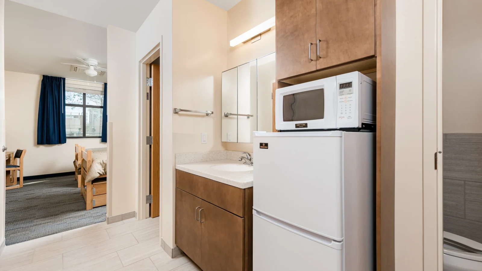 Unit kitchenette and vanity with wood cabinetry, a microwave and refrigerator, and a view through to the bedroom.
