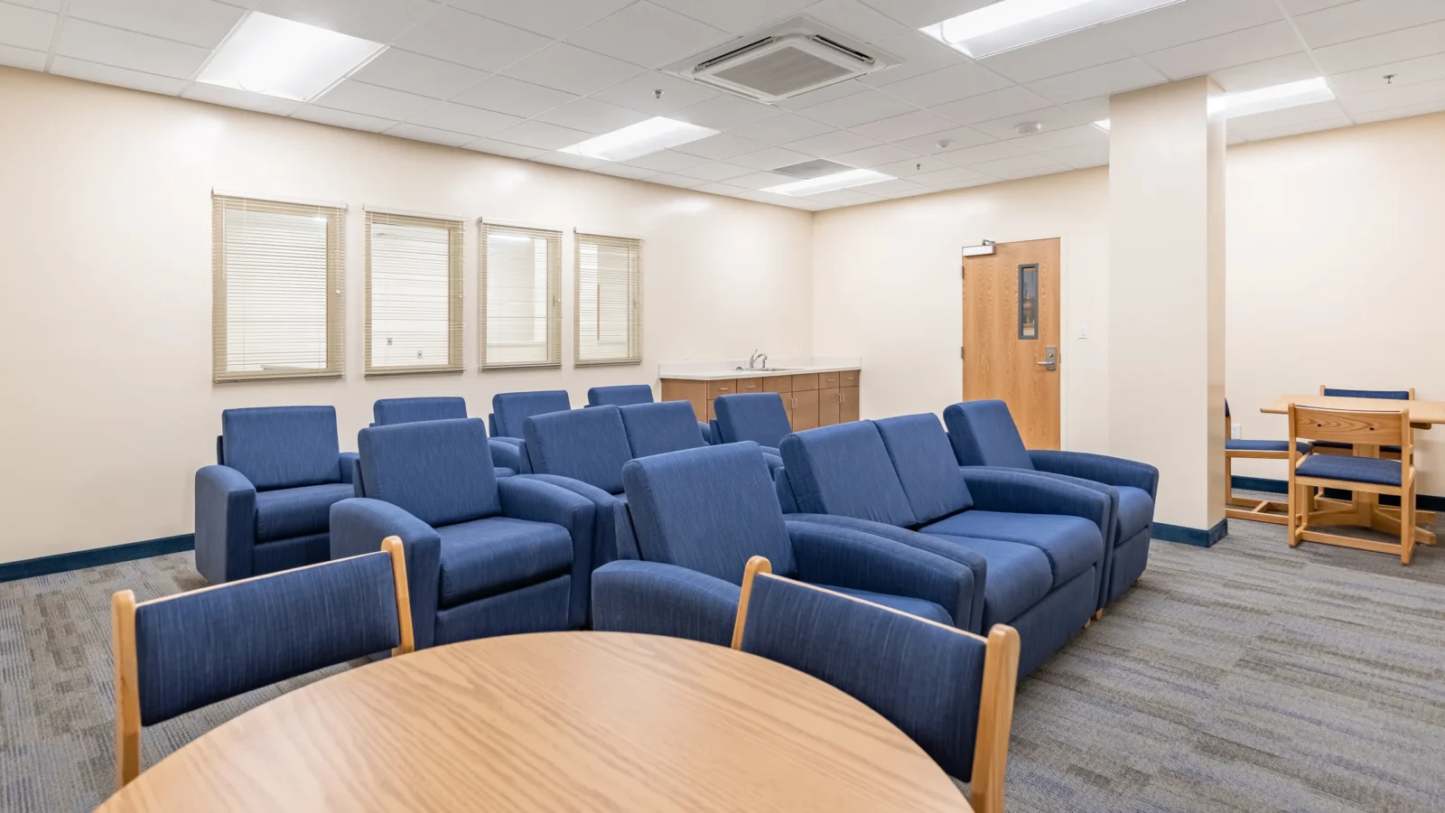 Residents' lounge with rows of blue upholstered seating, a round oak table, a kitchenette counter and clerestory windows.