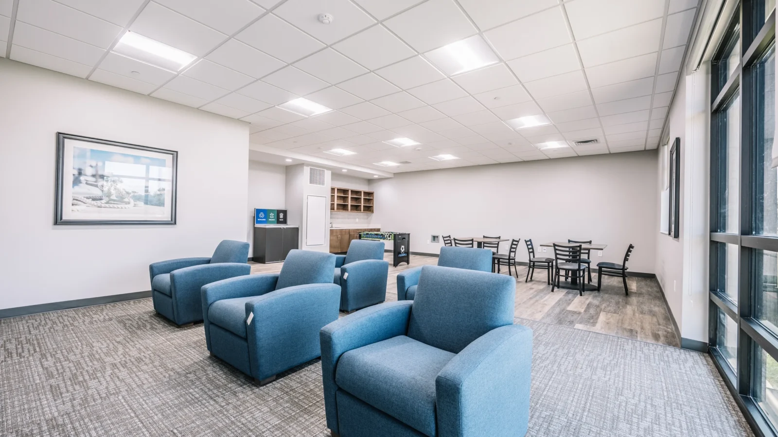 Residents' lounge with blue club chairs on carpet tile, a game and dining area on wood-look flooring, and full-height windows.