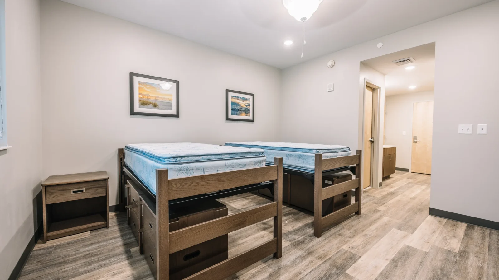 Two-person room with oak beds over storage drawers, nightstand, wood-look resilient flooring and an entry hall to the shared bath.