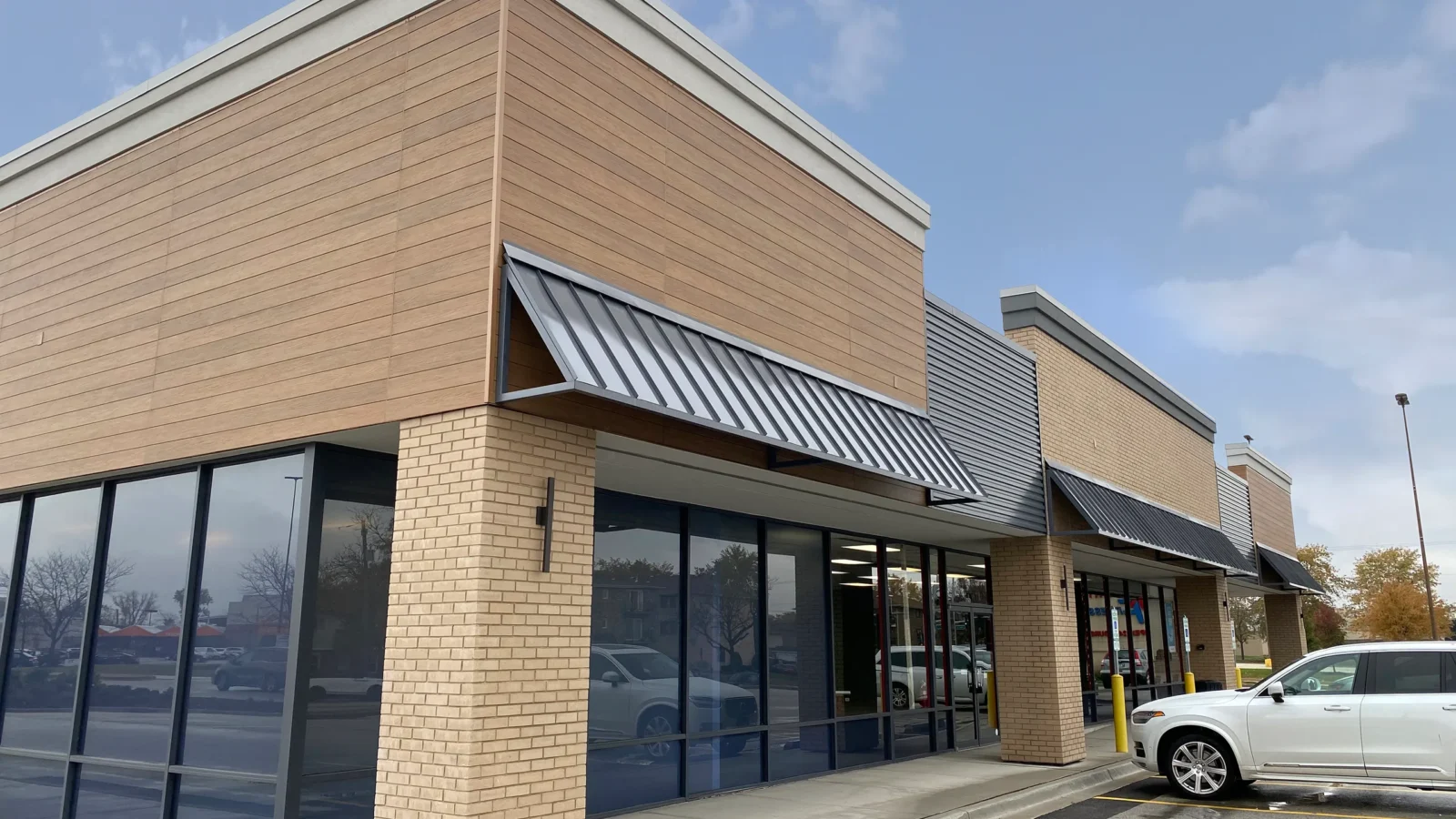 Close corner view of a remodeled building with wood-look siding, a tan brick column, angled metal awning and tall storefront glass.