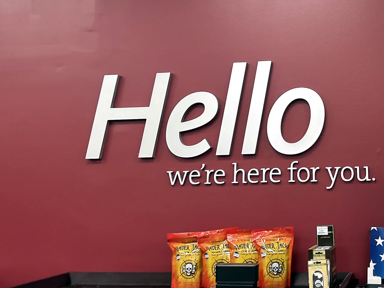 Shaw AFB Express Image Update 2.0 feature wall, white dimensional Hello, we're here for you lettering on a deep red wall above a dark wood display case.