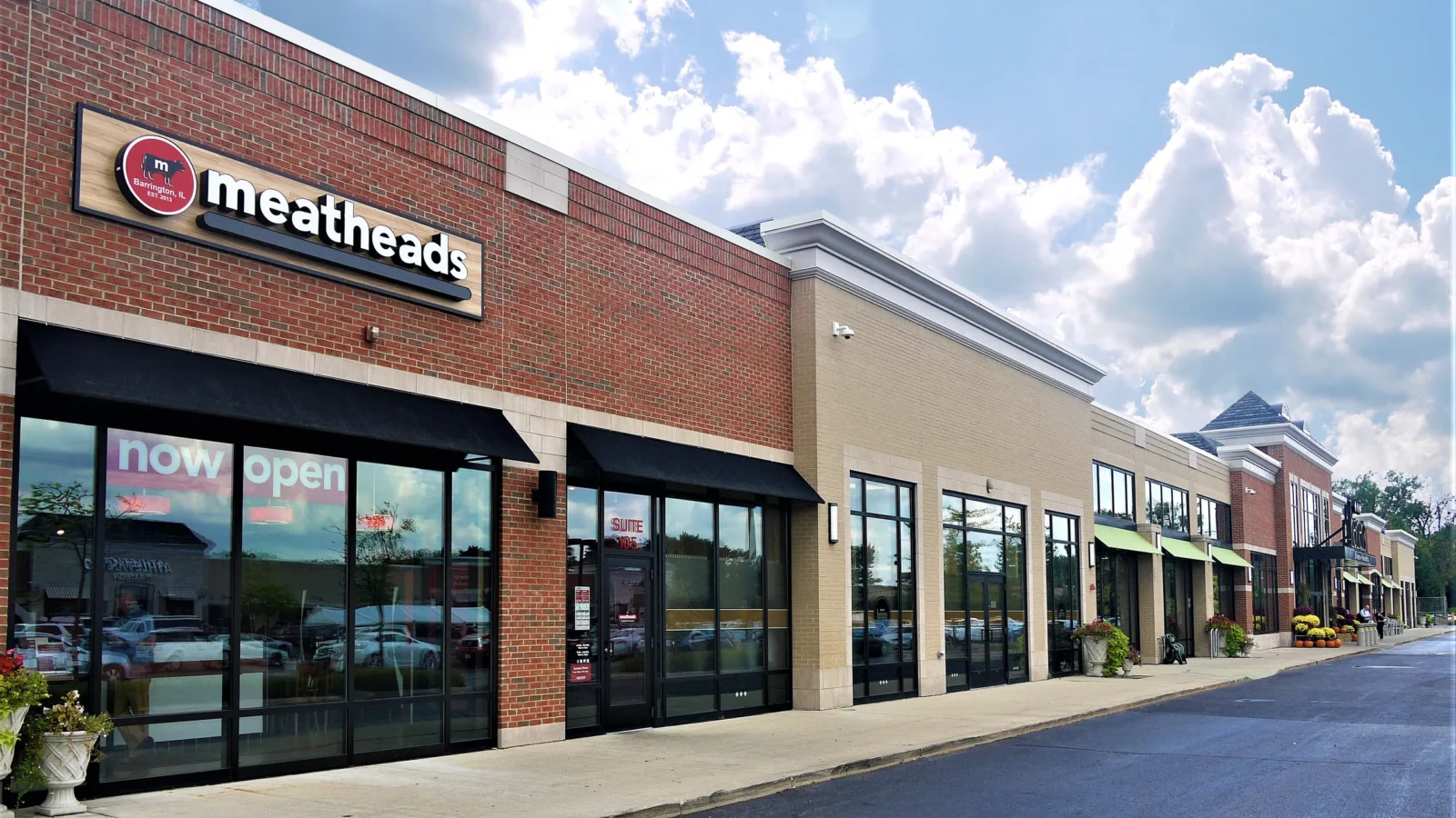 Shops at Flint Creek south shops: red and tan brick storefronts with limestone trim, black awnings and a long run of glass tenant fronts.