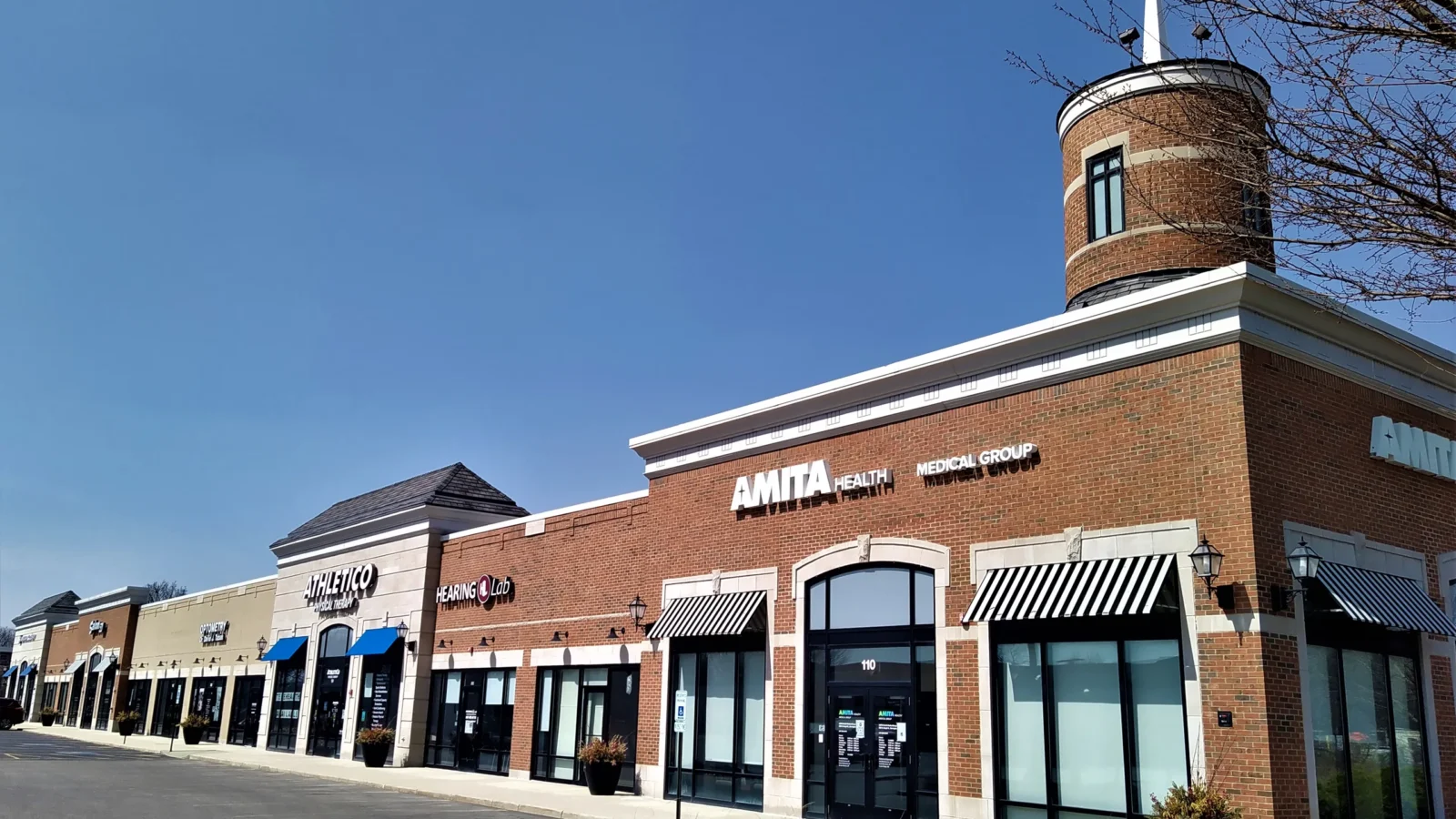 North shops with a round brick tower and white spire at the corner, striped awnings, arched entry and a row of tenant storefronts.