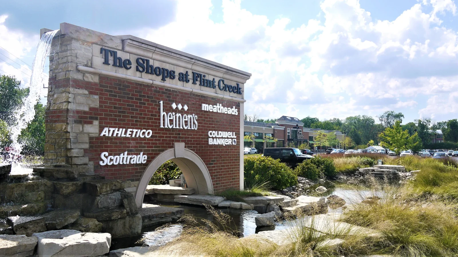 Brick and limestone monument sign for The Shops at Flint Creek with an arched opening over a stone waterfall and planted creek.