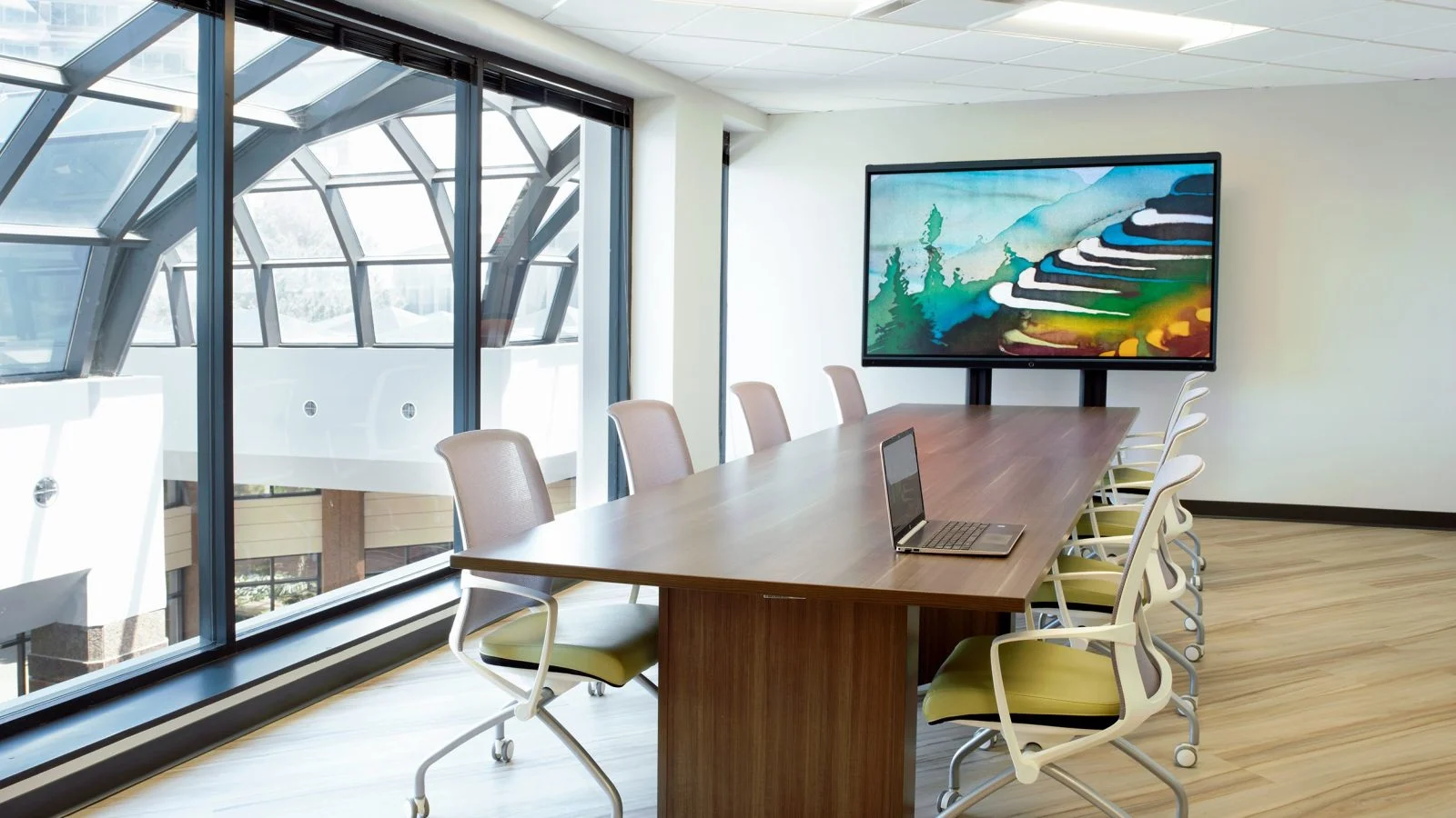 A boardroom table in dark timber set against a curved glazed wall, with a wall-mounted display showing artwork and pale timber flooring underfoot.