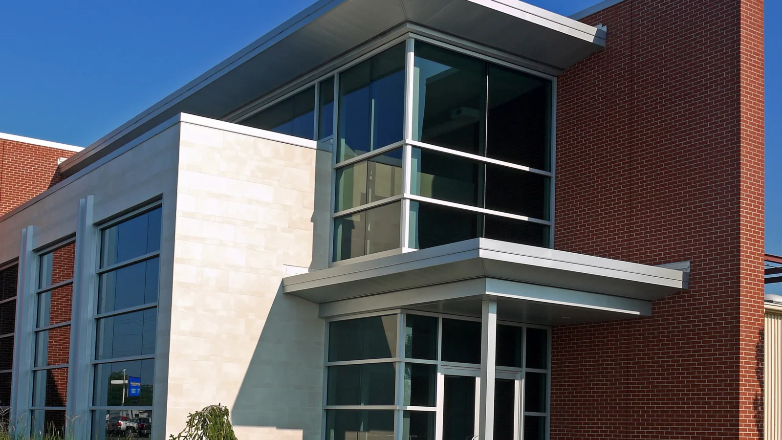 Glass entry under a cantilevered metal canopy, framed by a cream stone volume and a tall red brick wall against deep blue sky.
