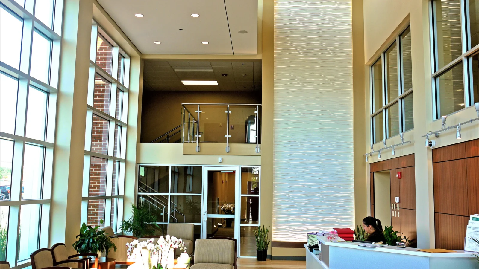 Two-story lobby with tall windows, a backlit sculpted wave wall, glass-railed overlook from the second floor and a white reception desk.