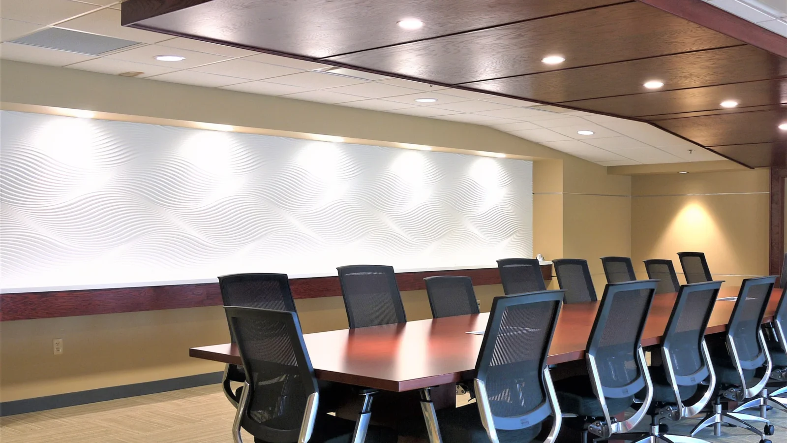 Boardroom with a long cherry table, mesh chairs, a dark wood ceiling soffit and a lit white wave-textured feature wall.