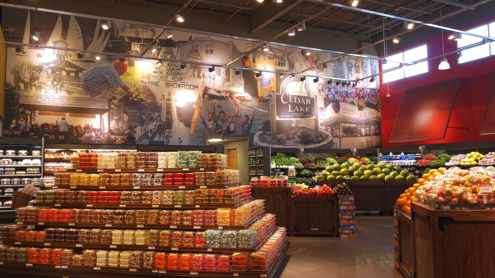 Produce department with wood display bins and a tiered candy table beneath a large historic Cedar Lake photo mural.
