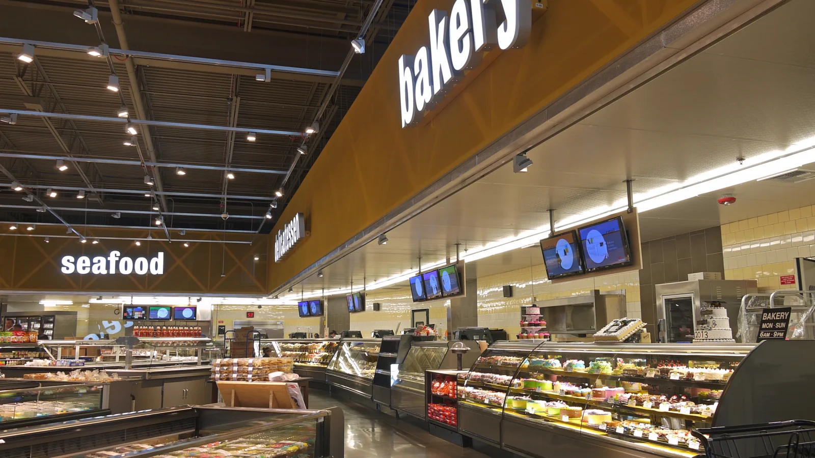 Service aisle with lit seafood, deli and bakery signs on a wood-toned fascia above curved glass display cases and a polished floor.