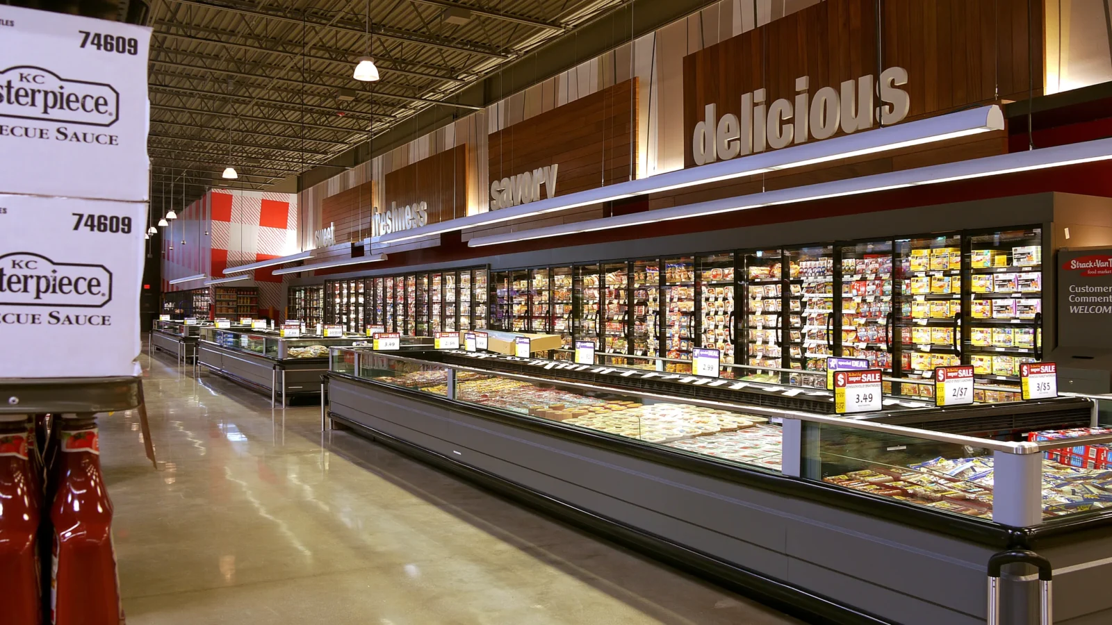 Long perimeter aisle of refrigerated cases under dark wood panels lettered savory and delicious, with an open ceiling above.