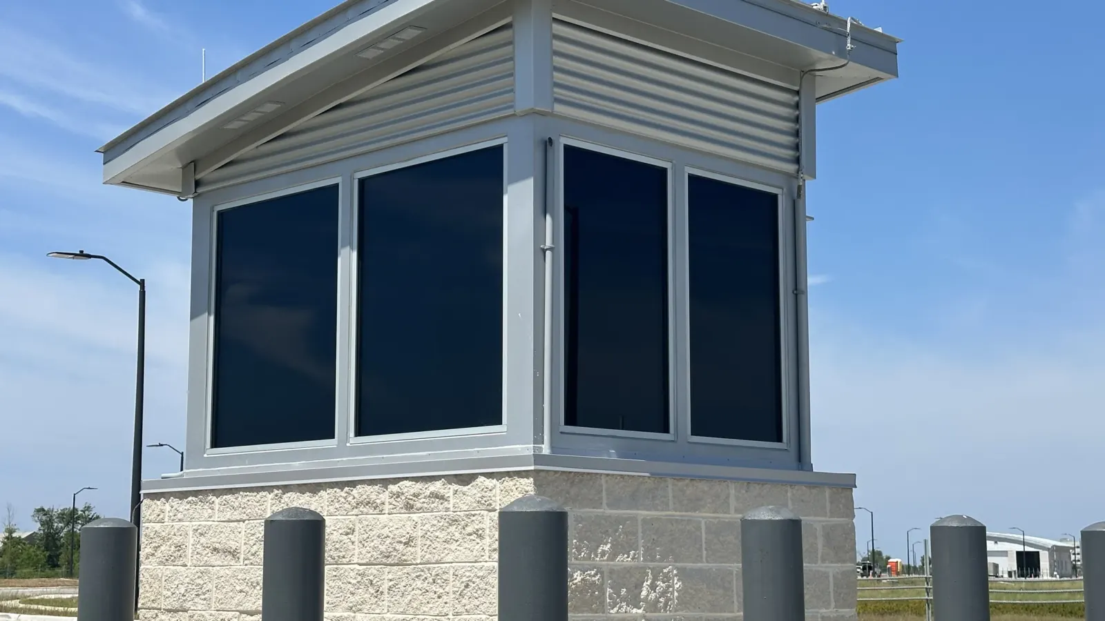 The glazed guard booth at the Tyndall AFB commercial gate, on a split-face block base behind steel bollards, under a deep metal roof.