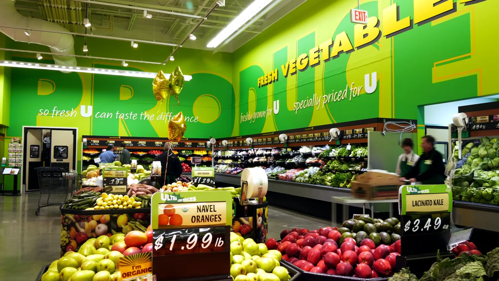 Ultra Grocery produce department under a bright green supergraphic wall reading Produce and Fresh Vegetables, with fruit displays in front.