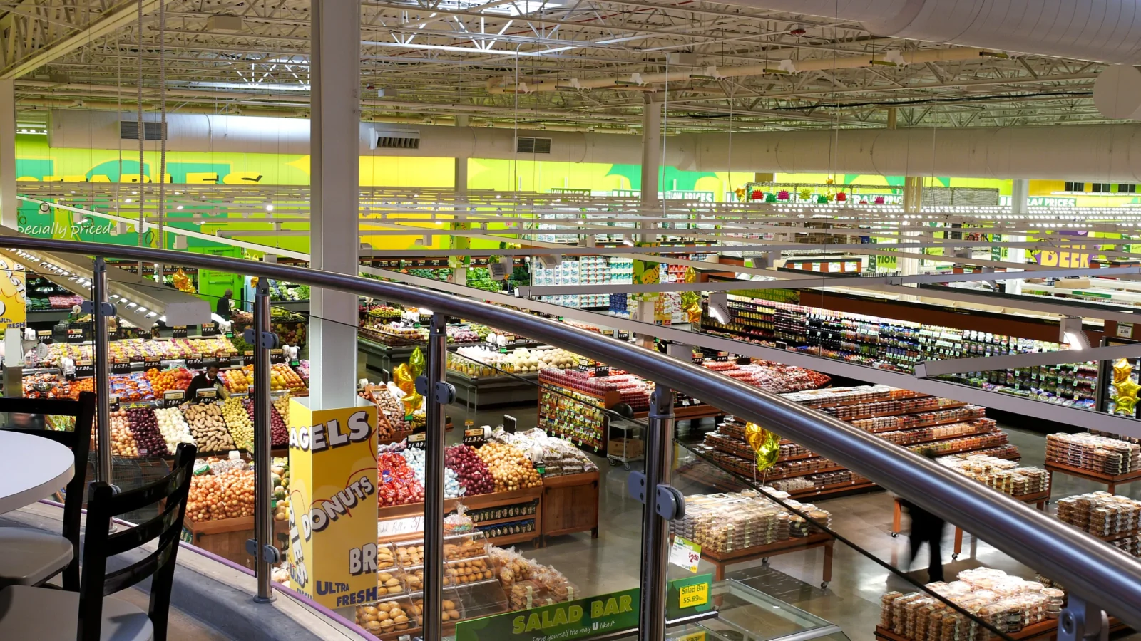 View from the glass-railed mezzanine across the sales floor, showing produce tables, bakery displays and a salad bar under the open ceiling.
