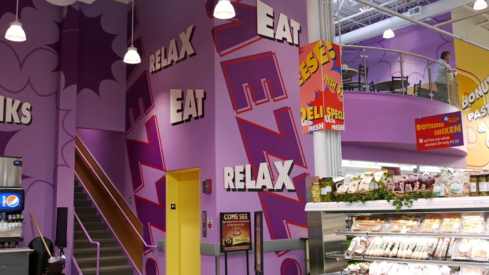 Purple cafe tower lettered Eat and Relax, with a stair to the mezzanine seating area and a yellow door beside the deli cases.