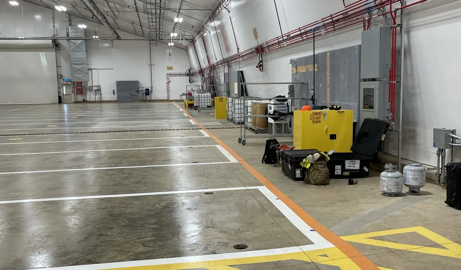 Along the hangar's service wall: yellow hatched safety markings and an orange walkway line on the polished floor, with electrical panels, piping and a flammables cabinet against the white membrane wall.